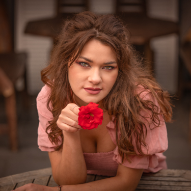 Portrait of a woman with long brown hair in a pink dress, leaning toward the camera over a garden table, holding a red flower beneath her chin with a subtle smile.