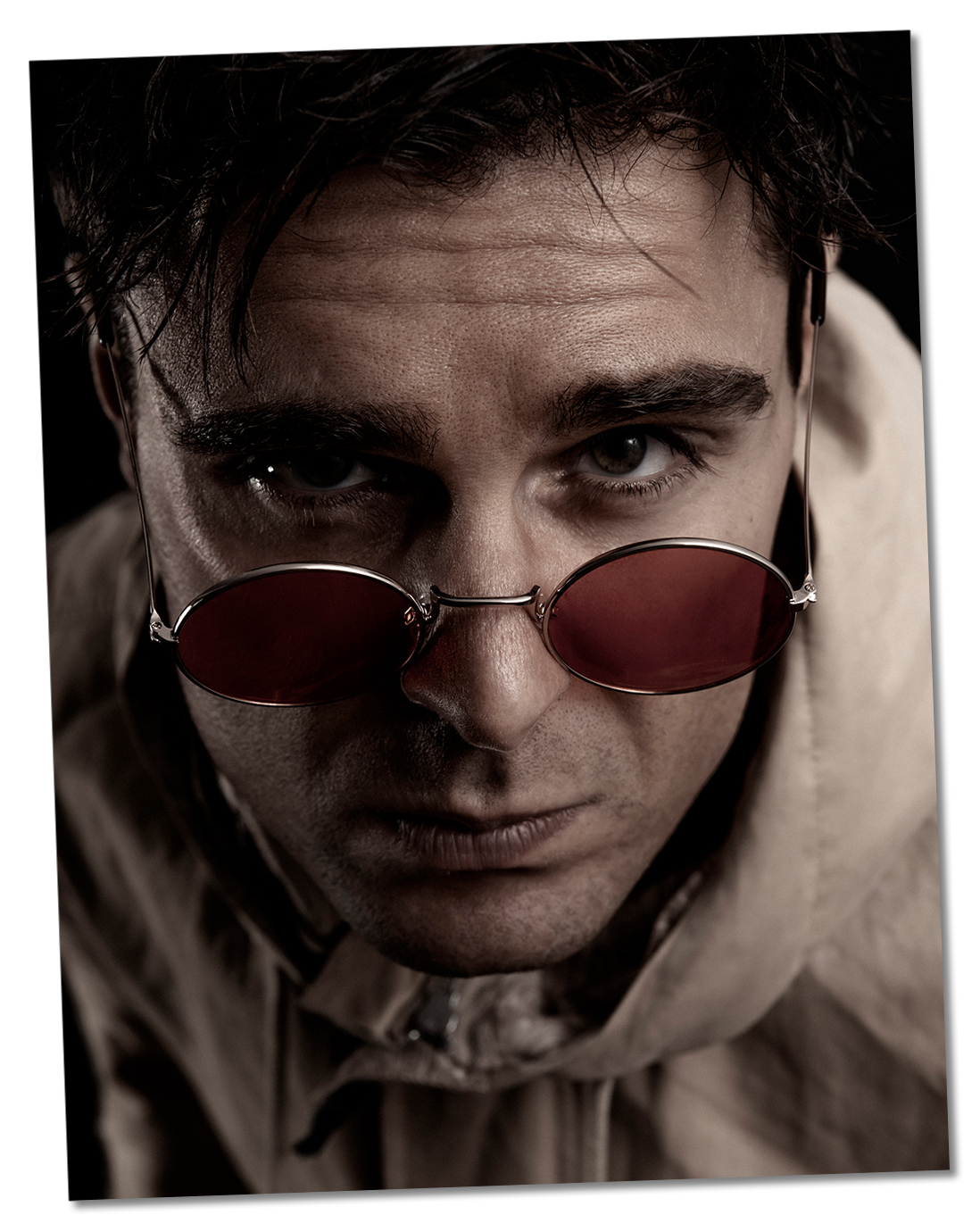 Close-up portrait of Ed Akay leaning slightly forward, wearing a light-coloured hoodie and dark red tinted round glasses perched low on his nose, looking directly at the camera with a confident, engaging expression. Cropped just above his fringe, showing part of the hoodie and his face.