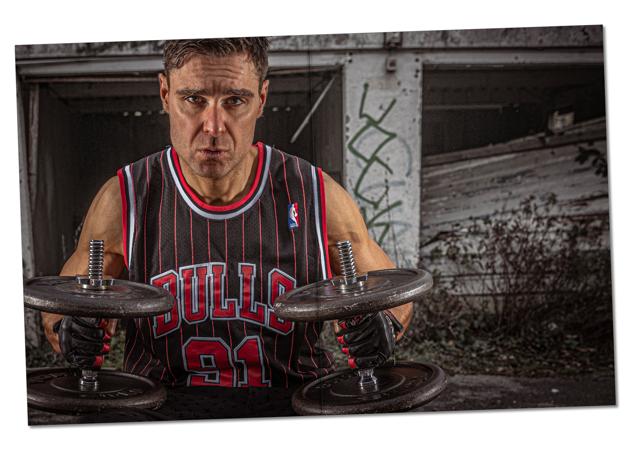 Ed Akay wearing a black and red Chicago Bulls vest with NBA emblem and weightlifting gloves, holding two dumbbells while staring blankly at the camera. Shot from waist to just above his fringe, he stands in front of a subdued, grainy background of old garage units—one without a door, the other with a door pushed in and resting on a pile of debris, with graffiti visible on the column between them. Strong lighting highlights Ed’s form and the colors of his vest.