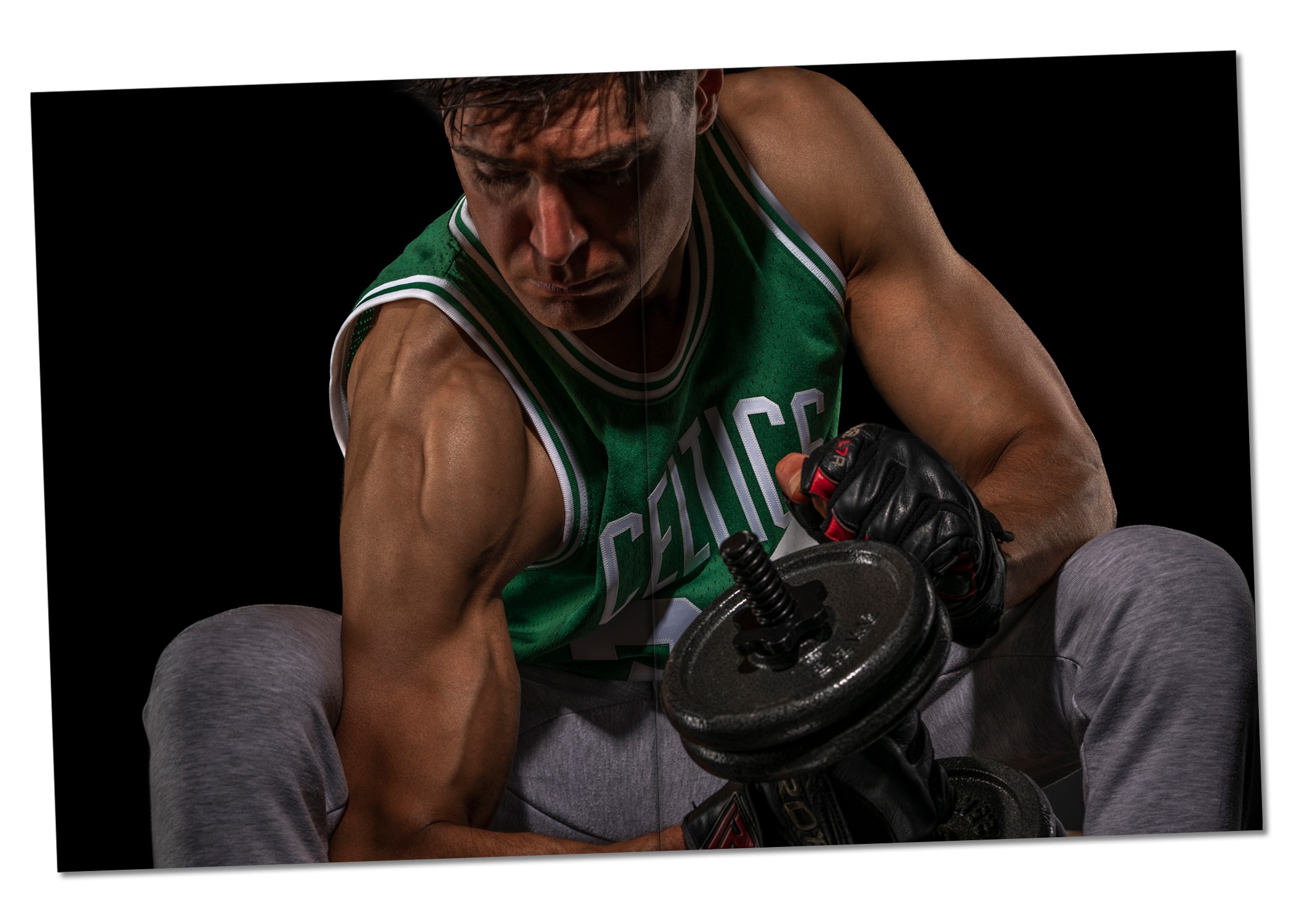 Ed Akay in studio on black backdrop, seated and leaning forward, curling a dumbbell between his knees, wearing a green Celtics vest and weightlifting gloves, focused expression, dramatic lighting highlighting muscles and posture.