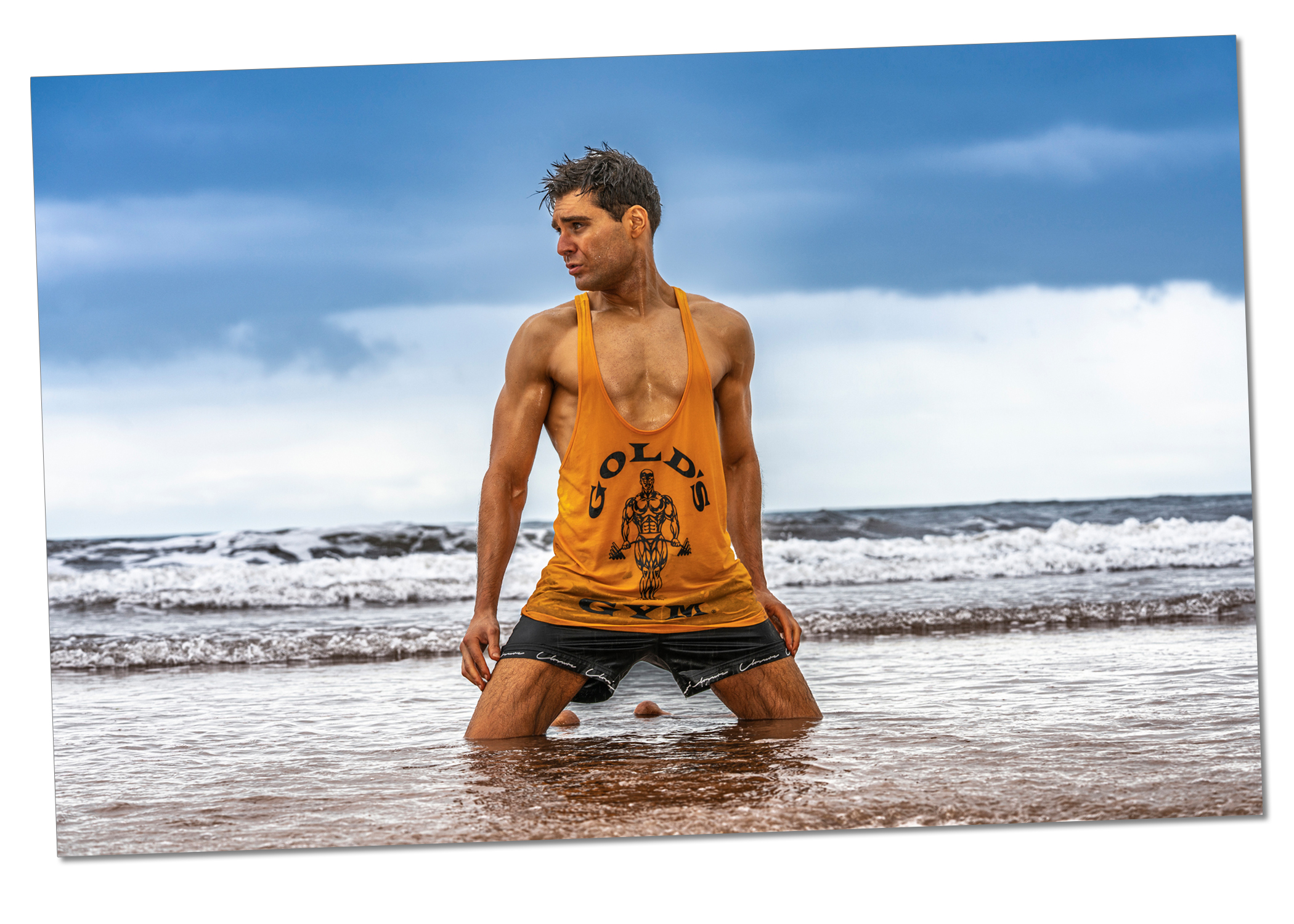 Page 36-37 from From Darkness, showing Ed Akay kneeling in the sea, water reaching halfway up his thighs. He wears a bright orange Gold’s Gym vest and dark shorts, with torso straight, arms at his sides, and head turned to the side. The horizon sits just below centre, sky partly blue with clouds near the horizon. Lighting balances Ed against the slightly overcast background, showing him clearly as the central focal point.