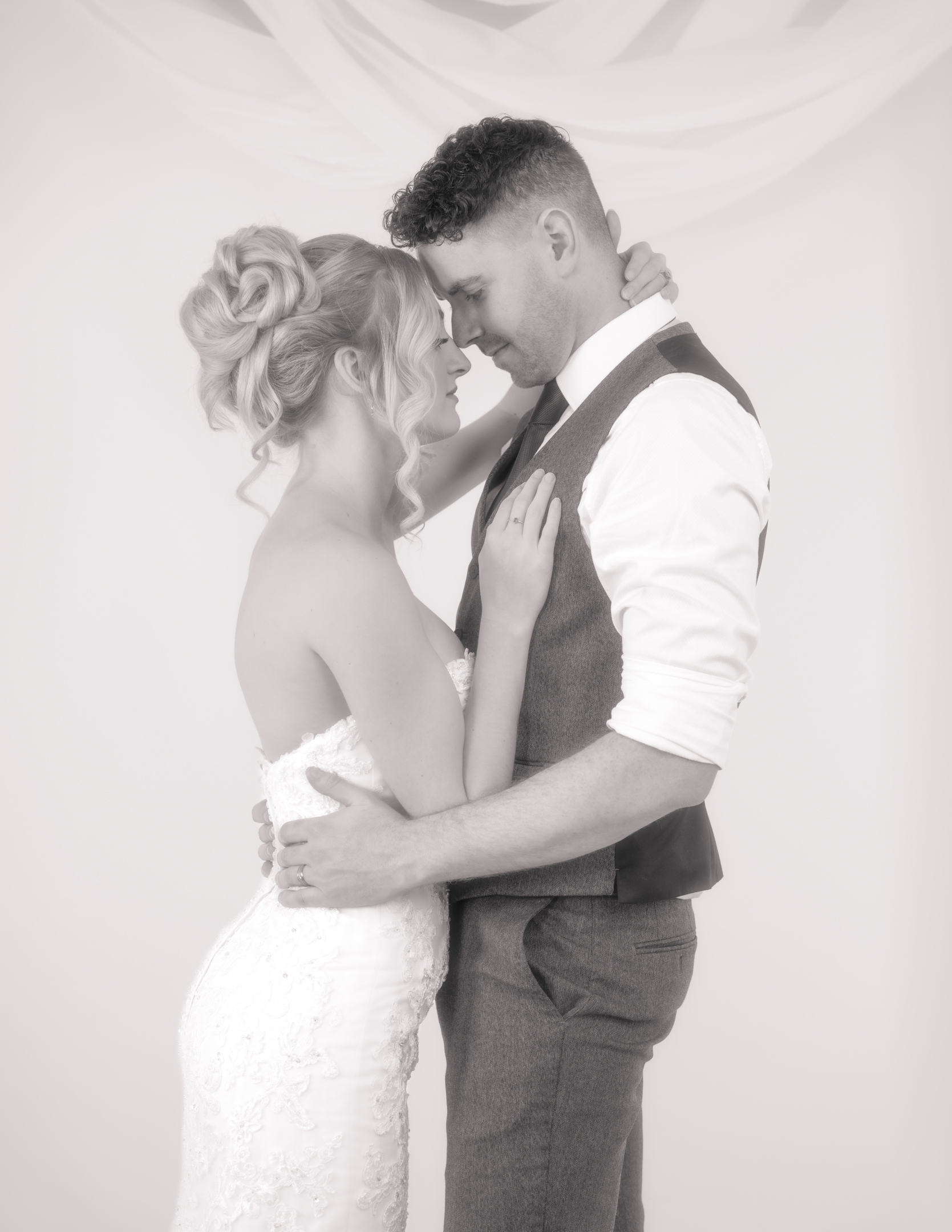 Orton effect high-key wedding portrait of bride and groom embracing; groom holds bride’s waist, bride has hand on chest and behind his neck, noses nearly touching, soft and gentle.