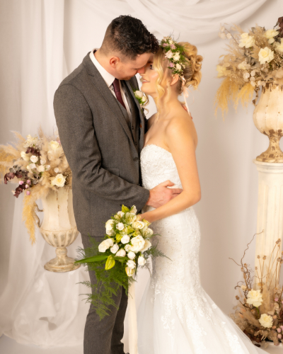 Colour wedding image of bride and groom almost kissing; bride smiling, holding bouquet with flowers in her hair, groom wearing buttonhole, white drapery and floral background.
