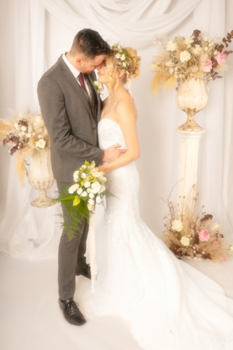 Light, airy wedding portrait with Orton effect; bride and groom facing each other, bride’s dress trailing on floor, bouquet in hand, white floral drapery behind.