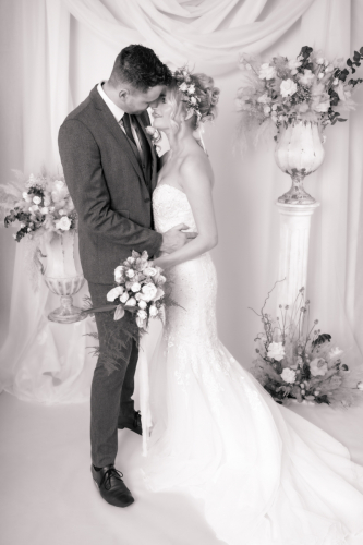 High-contrast monotone wedding image of bride and groom embracing; groom nuzzles bride’s hair with flowers and drapery in the background.