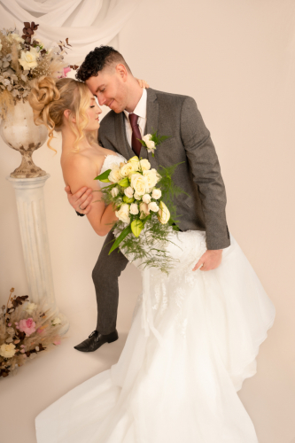 Bride and groom in playful tango-like pose; groom holds bride’s leg against his hip as she leans back, bouquet in hand, eyes closed, both smiling adoringly.