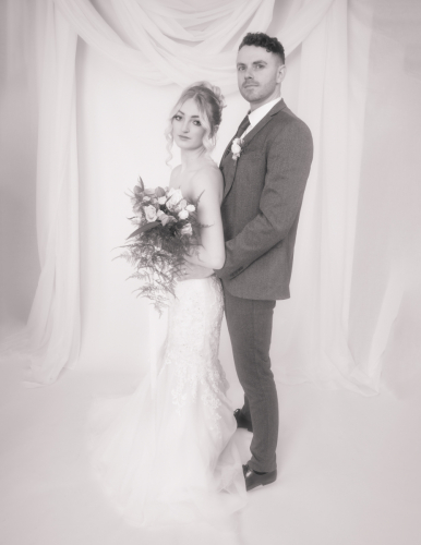 High-key, light Orton effect monotone portrait of bride and groom side-on; bride’s back against groom’s front, holding bouquet, white drapery behind, full-length dress visible.