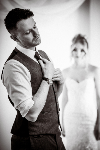 Cinematic high-contrast monotone wedding image; groom tightening tie in foreground, bride softly blurred in background, creating depth and grainy texture.