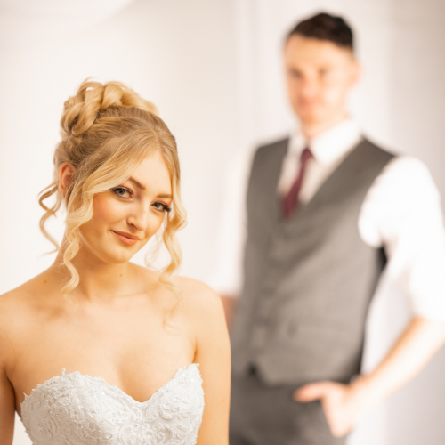 Colour wedding portrait focusing on bride’s bust and shoulders in foreground; she leans head to side, smiling gently, groom blurred in background, white backdrop.