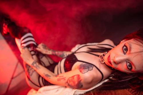 Model lying on back, legs raised, adjusting white stockings with black stripes, wearing Killstar lingerie, dramatic red smoke backdrop.