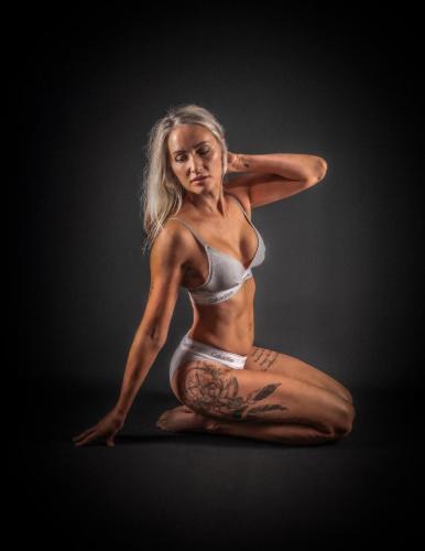 Stylish lingerie photoshoot with model kneeling sideways in Calvin Klein set, black backdrop emphasizing elegance and form.