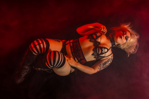 Blonde tattooed model lying on back with knees bent, wearing black skull-and-crossbones lingerie, black and white striped stockings, and knee-high black boots, shot from above with moody red accent lighting, Halloween-inspired style.