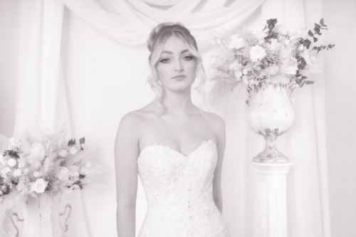 High-key, wide landscape Orton effect portrait of bride standing waist-up, straight-faced, looking at camera, white drapery and floral accents behind her.
