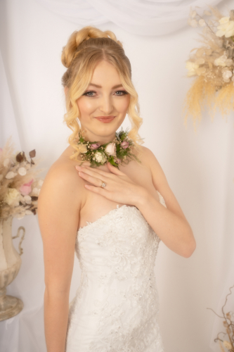 Colour high-key Orton effect close-up of bride smiling, hand on chest, floral necklace visible, subtle flowers in background.