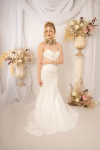 Light Orton effect full-length portrait of bride, left arm across waist showing wedding ring, floral necklace around neck, white drapery and urns of flowers in background.