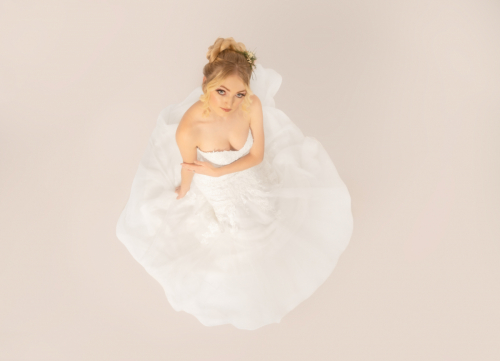 Top-down colour portrait of bride kneeling on floor, dress splayed out around her, looking up at camera, off-white floor emphasizing geometric dress shape.
