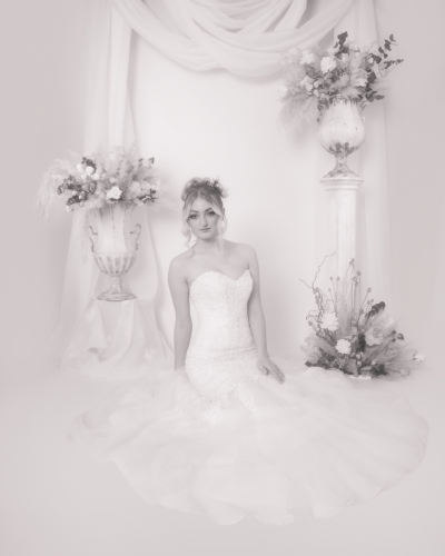 High-key monotone with pink tint and Autumn effect, bride kneeling on floor, viewed from front, dress spread around her, white drapery and floral urns in background.