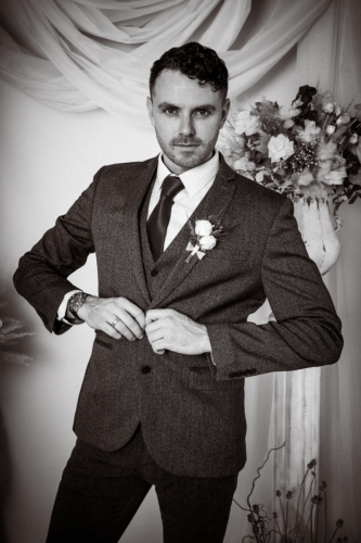 Cinematic dark portrait of groom buttoning jacket, elbows out, serious expression, flowers in urns and drapery behind, grainy texture, punchy contrast.
