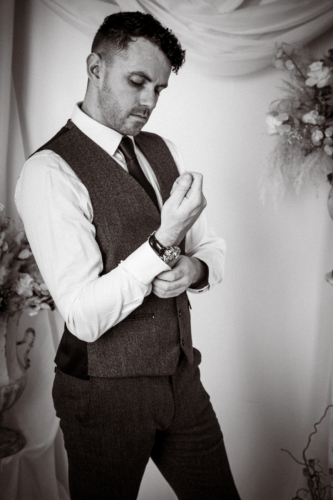 Cinematic dark portrait of groom doing up cufflink, standing sideways to camera, floral urns and drapery behind, high contrast with slight grain.