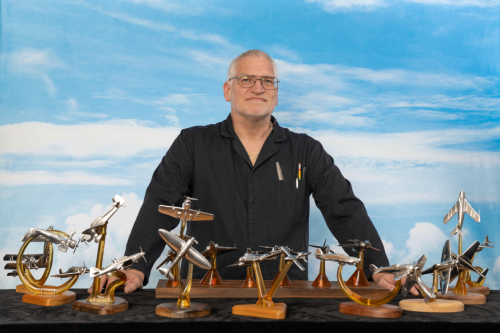 Robert Millis standing against a blue sky with his aluminium aerial combat sculptures displayed on a table.